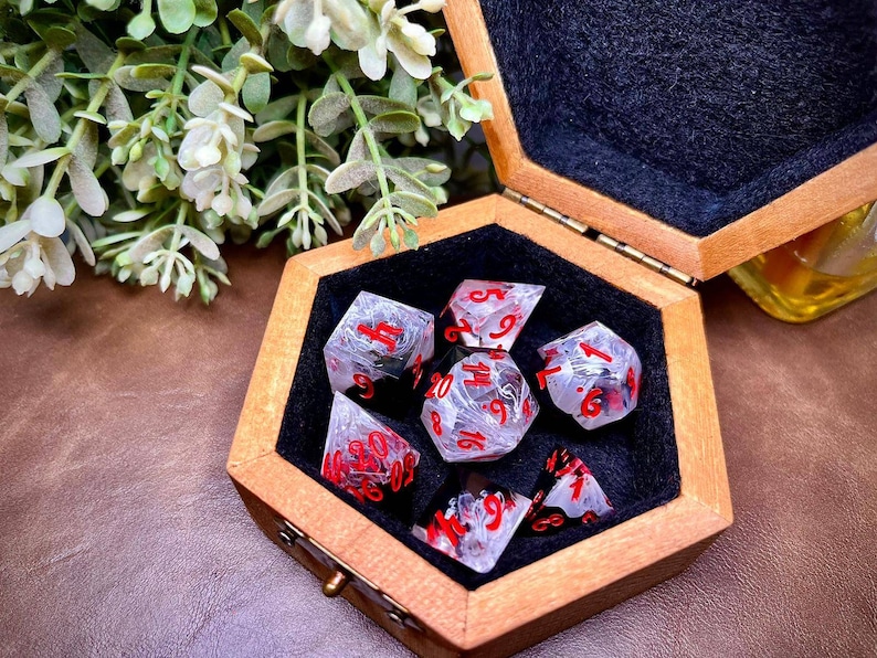 May include: A set of seven clear resin dice with red numbering, presented in a wooden hexagonal box lined with black felt. The dice have a marbled effect, suitable for tabletop games.