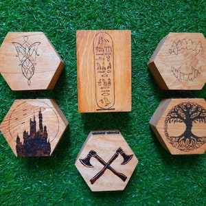 May include: A selection of six wooden, hexagonal boxes with different designs. The designs include a Celtic knot, Egyptian hieroglyphs, fangs, a castle silhouette, a tree of life, and crossed axes. The boxes are made of light brown wood.