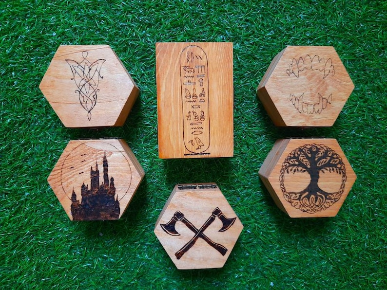 May include: A selection of six wooden, hexagonal boxes with different designs. The designs include a Celtic knot, Egyptian hieroglyphs, fangs, a castle silhouette, a tree of life, and crossed axes. The boxes are made of light brown wood.