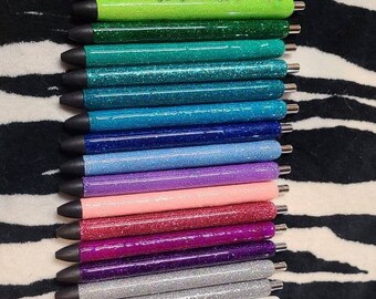 Può includere: Una collezione di penne glitterate colorate, tra cui rosso, arancione, verde e blu. Ogni penna ha la punta nera e dettagli argentati. Le penne sono disposte su uno sfondo a righe zebrate in bianco e nero.
