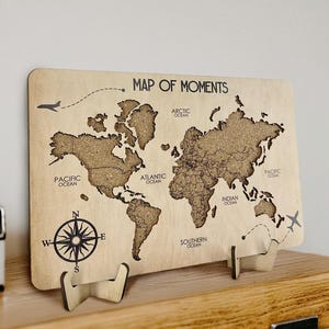 May include: A wooden world map with the text "MAP OF MOMENTS" and a compass rose. The map is supported by two wooden stands and is displayed on a wooden shelf. The map features the continents and oceans.