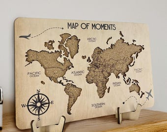 May include: A wooden world map with the text "MAP OF MOMENTS" and a compass rose. The map is supported by two wooden stands and is displayed on a wooden shelf. The map features the continents and oceans.