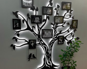 Puede incluir: Una silueta de árbol de metal negro con un contorno de luz LED blanca. El árbol tiene ramas que sostienen 10 marcos de fotos negros con fotos de familia. El árbol está montado en una pared blanca. Una pequeña planta en maceta se encuentra en la esquina inferior derecha de la imagen.