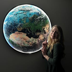 Op de afbeelding: Een ronde, verlichte wandkunst die de aarde afbeeldt. Het kunstwerk toont een gedetailleerde kaart van de continenten en oceanen, met een heldere, gloeiende rand. De kleuren zijn blauw, groen, bruin en wit, wat een driedimensionaal effect creëert.