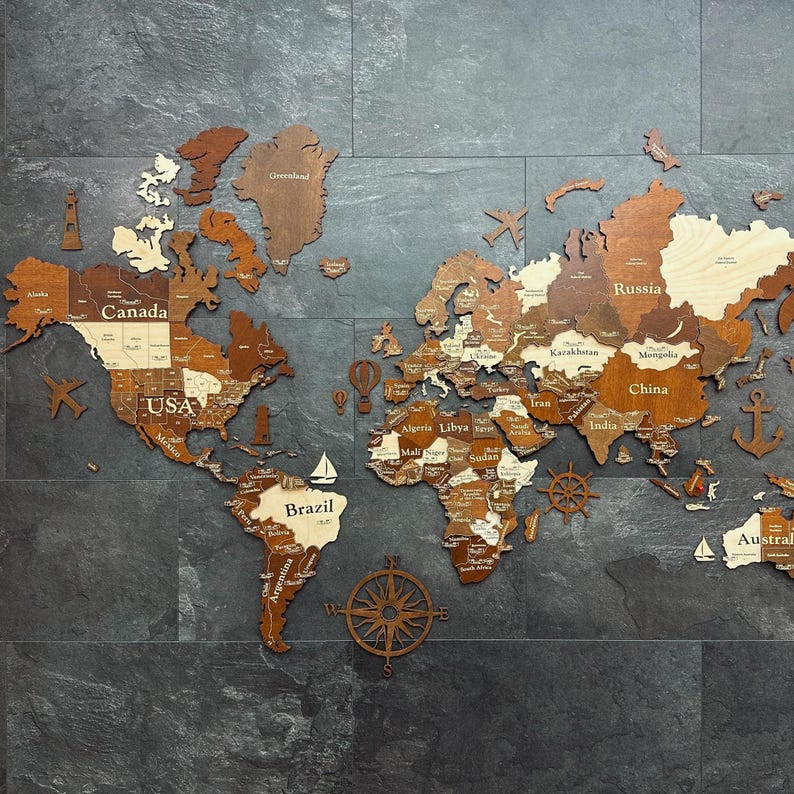 May include: A wooden world map with countries and continents in varying shades of brown and tan. Decorative nautical elements like a compass rose, sailboats, and an anchor are included. The map is mounted on a dark gray tiled surface.