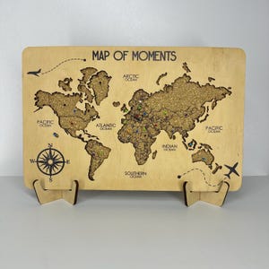 May include: A wooden world map with the text "MAP OF MOMENTS" and ocean labels. The map features cork detailing for the continents and colorful pins. A compass rose and airplane illustrations are also present.