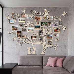 Puede incluir: Una decoraci&oacute;n de pared de madera con forma de &aacute;rbol con ramas y hojas. El &aacute;rbol est&aacute; decorado con m&uacute;ltiples marcos de fotos, cada uno con una imagen diferente. El &aacute;rbol tambi&eacute;n est&aacute; decorado con peque&ntilde;os recortes de madera de mariposas, p&aacute;jaros y b&uacute;hos. El &aacute;rbol est&aacute; montado en una pared gris, y hay un sof&aacute; gris en primer plano.