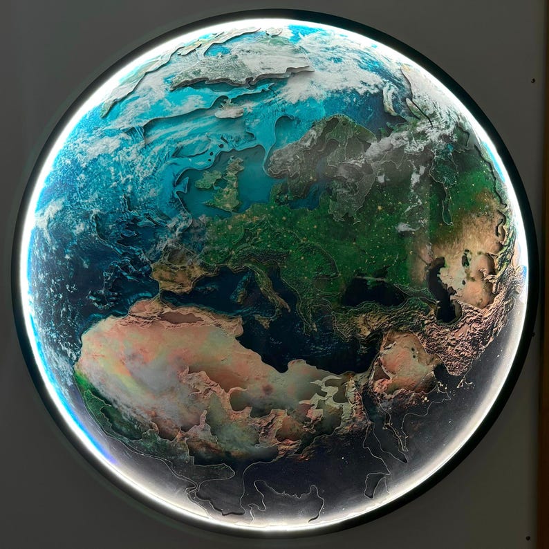 May include: A round, illuminated Earth map with a detailed relief, showcasing continents and oceans in shades of green, blue, and brown. The map is encased in a black frame with a bright white light around the edge, creating a glowing effect.