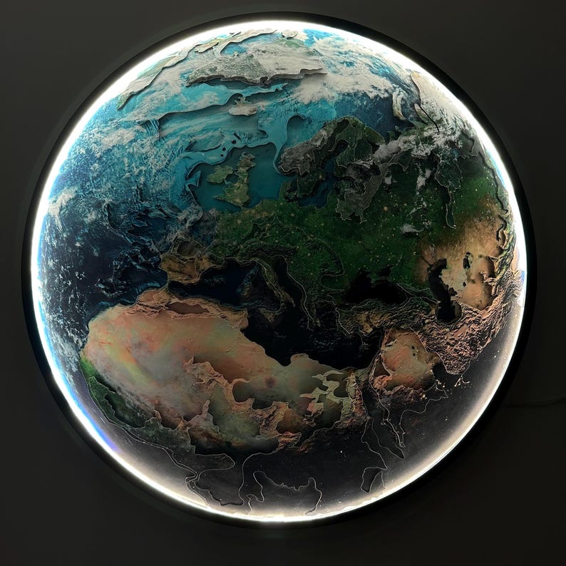 May include: A round, illuminated wall art piece depicting a detailed relief map of Earth. The artwork features a black frame with a bright white light outlining the globe. The continents are rendered in shades of green, brown, and tan, with blue oceans.