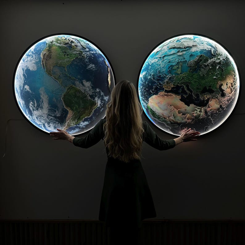 May include: Two illuminated Earth globes with detailed landmass and ocean features are mounted on a wall. A person stands between them, arms outstretched. The globes have a bright, glowing edge.