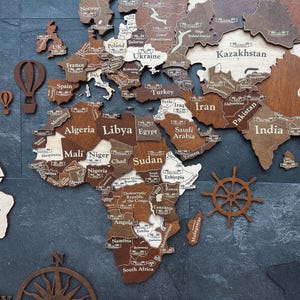 May include: A wooden world map with country names engraved in white. The map features varying shades of brown and tan, with decorative elements like a compass, hot air balloons, and a ship's wheel. The map is mounted on a dark gray surface.