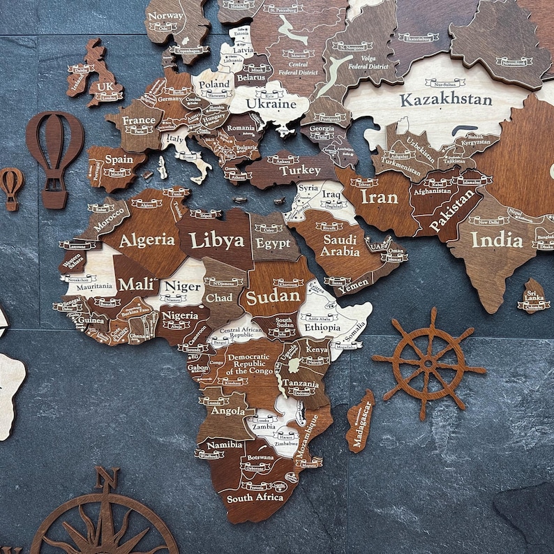May include: A wooden world map with country names engraved in white. The map features varying shades of brown and tan, with decorative elements like a compass, hot air balloons, and a ship's wheel. The map is mounted on a dark gray surface.