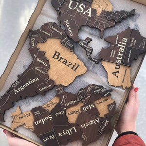 May include: A wooden world map puzzle with continents and countries labeled. The map is in a rectangular box, with the USA, Brazil, and Australia prominently displayed. The continents are in shades of brown and tan.