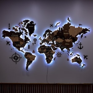 Könnte beinhalten: Eine Weltkarte aus Holz mit beschrifteten Ländern, beleuchtet mit blauen LED-Leuchten. Dekorative Ausschnitte von Flugzeugen, einem Kompass und einem Steuerrad sind enthalten. Die Karte ist an einer weißen Wand befestigt.