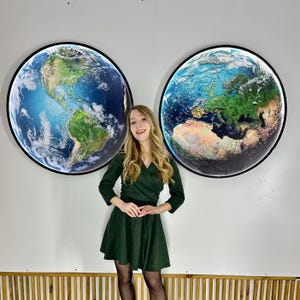 May include: Two large, round wall art pieces depicting Earth, with continents and oceans in vibrant colors, are displayed on a white wall. Each piece is framed in a black border. A person in a green dress stands in front of the artwork.