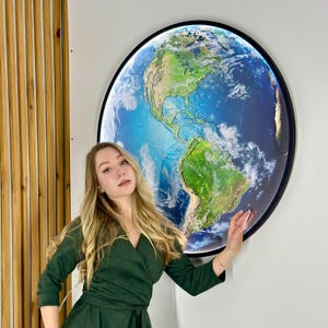 May include: A large, round illuminated wall art piece depicting a detailed map of Earth. The artwork features continents in shades of green and brown, surrounded by blue oceans and white clouds. The piece is framed in black and mounted on a white wall.