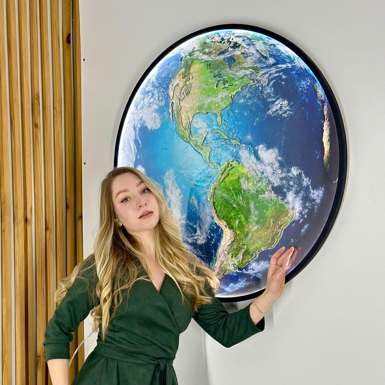 May include: A large, round illuminated wall art piece depicting a detailed map of Earth. The artwork features continents in shades of green and brown, surrounded by blue oceans and white clouds. The piece is framed in black and mounted on a white wall.