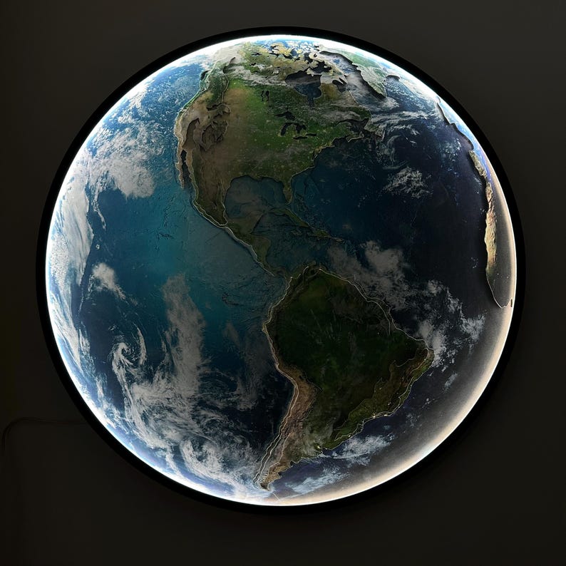May include: A round, illuminated wall art piece depicting Earth. The artwork shows the continents in shades of green and brown, surrounded by blue oceans and white clouds. The piece has a black frame and a bright, white light around the edge.