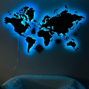 Könnte beinhalten: Eine schwarze Weltkarte aus Holz mit blauer LED-Hintergrundbeleuchtung, an einer Wand befestigt. Die Karte zeigt detaillierte Umrisse von Kontinenten und Ländern. Dekorative nautische Elemente, darunter ein Kompass und Anker, sind ebenfalls sichtbar.