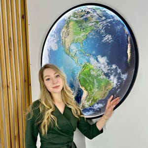 May include: A large, round wall art piece depicting a detailed map of Earth. The artwork features a black frame and is illuminated, showcasing the continents and oceans in vibrant colors. The piece is mounted on a white wall.