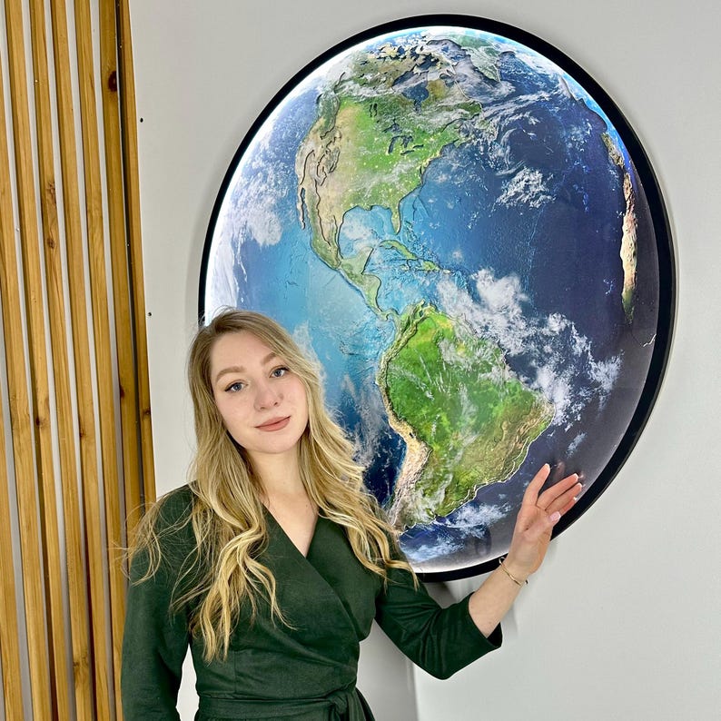 May include: A large, round wall art piece depicting a detailed map of Earth. The artwork features a black frame and is illuminated, showcasing the continents and oceans in vibrant colors. The piece is mounted on a white wall.