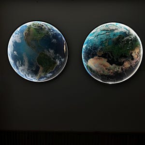 May include: Two illuminated, round wall art pieces depicting Earth. One shows the Americas, the other shows Europe, Africa, and Asia. The globes have a black border and a bright white light around the edge, creating a striking contrast against the dark background.