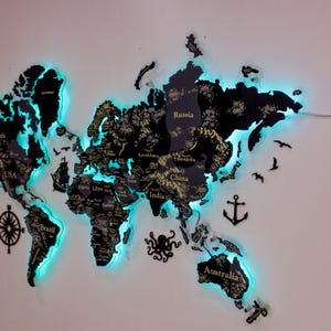 Puede incluir: Un mapa del mundo negro montado en la pared con etiquetas de texto de color dorado para países y continentes. El mapa cuenta con una retroiluminación LED cian brillante que crea un efecto de resplandor en los bordes. Se incluyen siluetas negras decorativas de un ancla, un pulpo y aves.