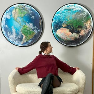 May include: Two illuminated circular wall art pieces depicting Earth from space. One shows the Americas, the other Europe and Africa. Each has a black frame and a bright white light around the edge. The art is mounted on a light gray wall.