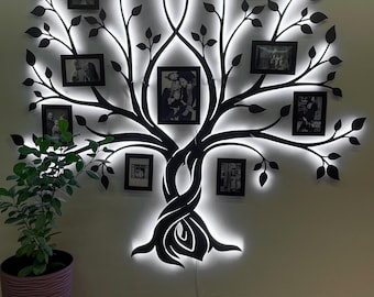 Puede incluir: Una silueta de árbol de metal negro con un contorno de luz LED blanca. El árbol tiene ramas que sostienen varios marcos de fotos negros. El árbol está montado en una pared blanca.