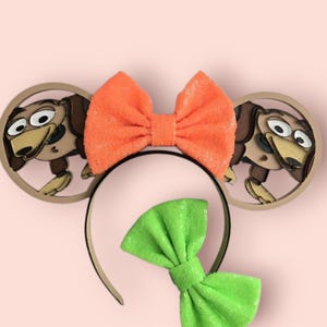 May include: A black headband with two circular ears featuring a cartoon dog design. An orange sequined bow is centered above the headband, and a green sequined bow is below it. The background is a solid light pink.