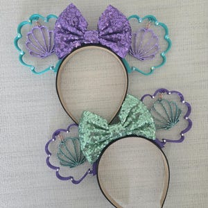 May include: Two pairs of purple and green sequined Minnie Mouse ears with a seashell design. The ears are decorated with a purple bow and a green bow.