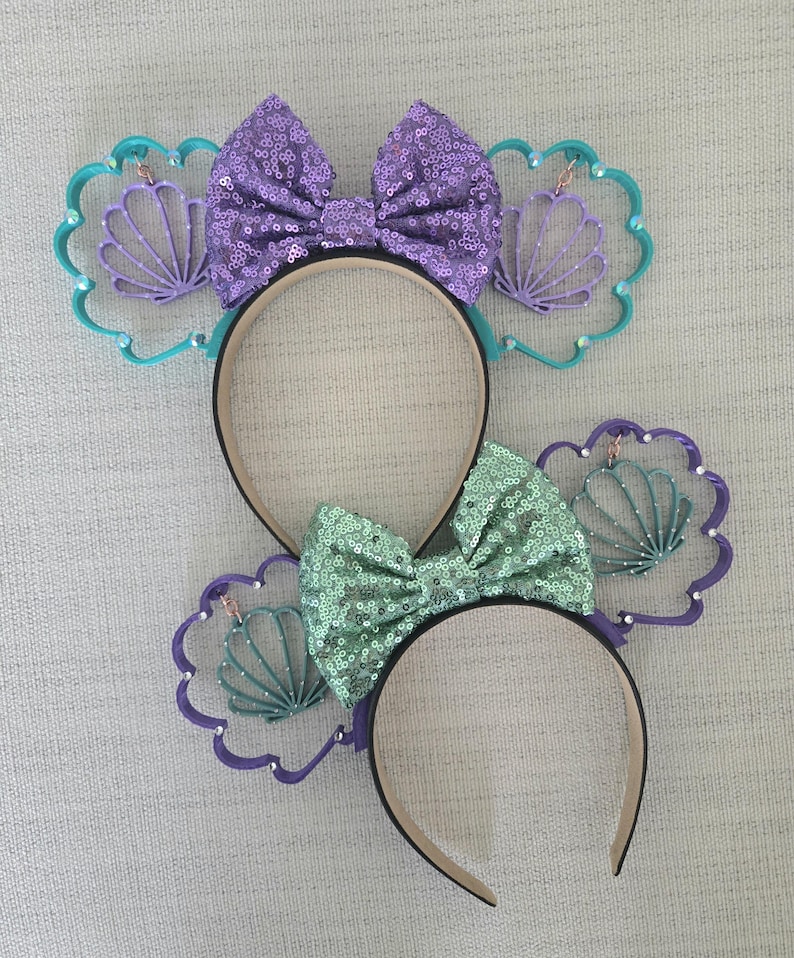 May include: Two pairs of purple and green sequined Minnie Mouse ears with a seashell design. The ears are decorated with a purple bow and a green bow.