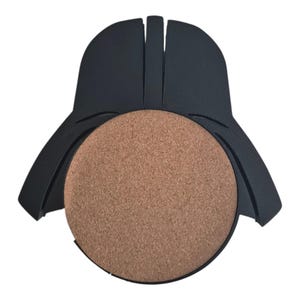 May include: A black plastic coaster holder shaped like a helmet with a circular brown cork insert in the center. The holder features a smooth matte finish and a distinct geometric design with a vertical ridge running down the top section.