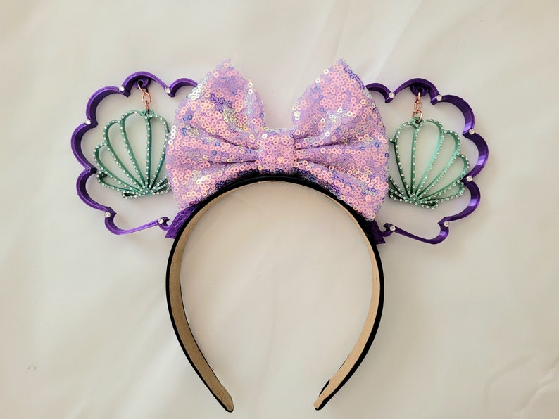 May include: Purple headband with a large lavender bow and two green seashell charms. The headband is decorated with rhinestones.