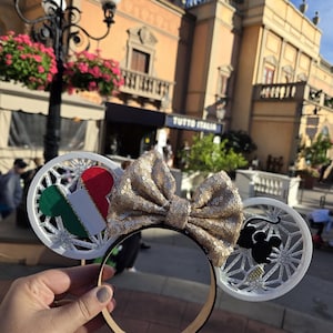 May include: White Minnie Mouse ears with a gold glitter bow. One ear features a white and silver snowflake design with a green, white, and red Italian flag. The other ear features a white and silver snowflake design with a black silhouette of Mickey Mouse.