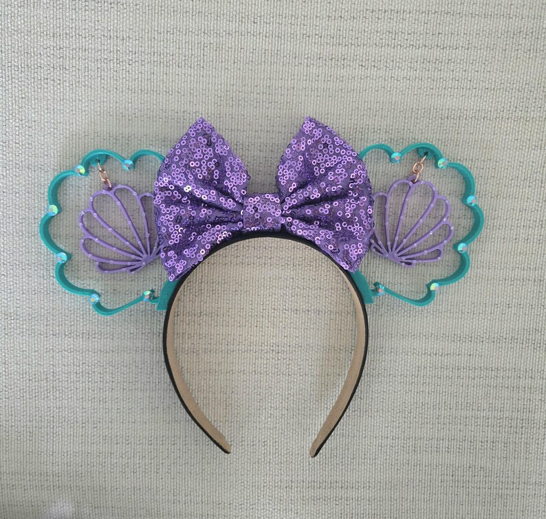 May include: Purple and teal Minnie Mouse ears with a purple sequined bow and a seashell design.