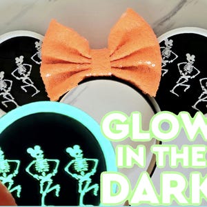 Pode incluir: Um par de orelhas de rato brancas com fundo preto e um desenho de três esqueletos dançando que brilha no escuro. As orelhas têm um laço laranja no topo. O texto "GLOW IN THE DARK" está escrito em letras verdes.