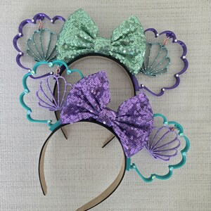 May include: Two pairs of purple and green mouse ears with a seashell design. The ears are decorated with a large bow and a smaller seashell charm.
