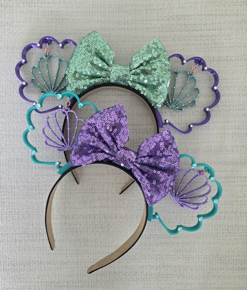 May include: Two pairs of purple and green mouse ears with a seashell design. The ears are decorated with a large bow and a smaller seashell charm.