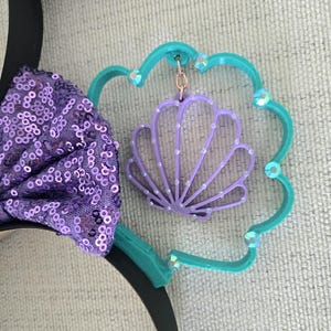 May include: Purple and teal Minnie Mouse ears with a purple seashell charm hanging from the teal ear. The seashell has a white pearl design.
