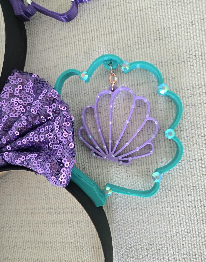 May include: Purple and teal Minnie Mouse ears with a purple seashell charm hanging from the teal ear. The seashell has a white pearl design.