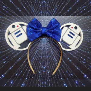 May include: White R2-D2 themed Minnie Mouse ears with a blue sequin bow. The ears are shaped like the iconic Star Wars character's head.