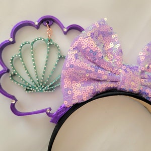 May include: Purple headband with a large iridescent purple bow and a purple seashell charm. The seashell charm has a green and white design and is attached to the headband with a silver chain.