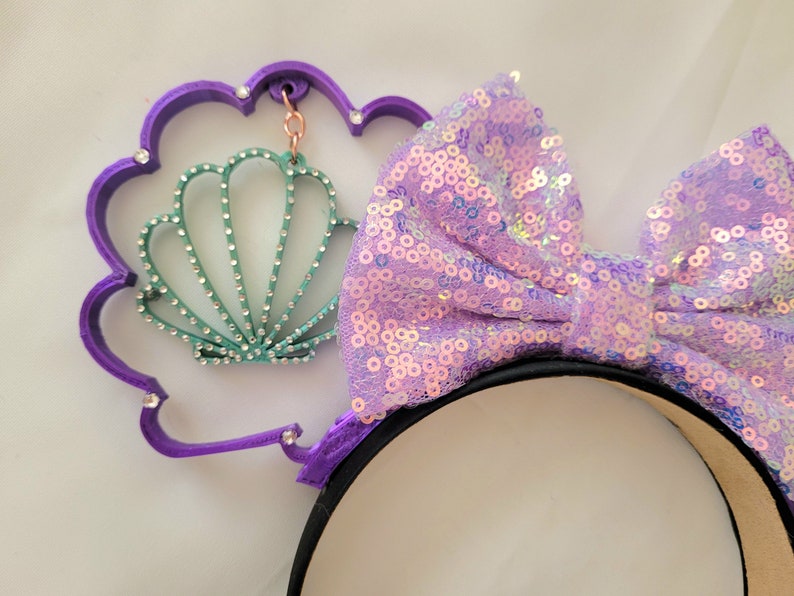 May include: Purple headband with a large iridescent purple bow and a purple seashell charm. The seashell charm has a green and white design and is attached to the headband with a silver chain.