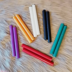 Könnte beinhalten: Satz mit acht dünnen Kerzen in verschiedenen Farben, darunter lila, orange, rot, grün, schwarz und weiß. Die Kerzen sind auf einer flauschigen, hellbraunen Oberfläche angeordnet.