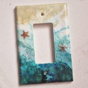 Peut inclure: Une plaque de prise de courant avec une scène de plage. La plaque est peinte avec des couleurs bleues, vertes et blanches pour ressembler à des vagues océaniques. Deux étoiles de mer dorées à paillettes sont sur la plaque.