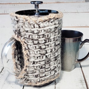 Peut inclure: Une housse en crochet gris et blanc pour une cafetière à piston. La housse est faite avec un fil épais et a un aspect rustique.