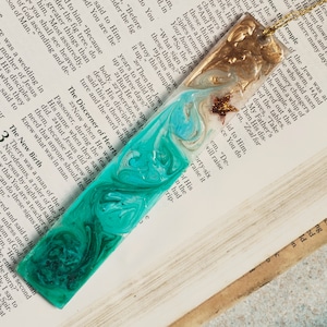 May include: Two handmade resin bookmarks with a marbled design. One bookmark is teal and blue with a gold glitter star. The other bookmark is gold and brown with a gold glitter star. The bookmarks are on top of an open book with text from the Bible.