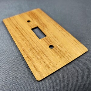 Könnte beinhalten: Eine Holz-Lichtschalterabdeckung mit einer rechteckigen Öffnung und zwei kreisförmigen Löchern. Die Abdeckung hat ein natürliches Holzmaserungsmuster und eine warme, hellbraune Farbe. Der Artikel ist rechteckig mit abgerundeten Ecken.
