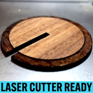 May include: A wooden, circular object with a dark brown outer ring and a lighter brown center. A black rectangular slot is cut through the center. The text "LASER CUTTER READY" is displayed in a blue box at the bottom.
